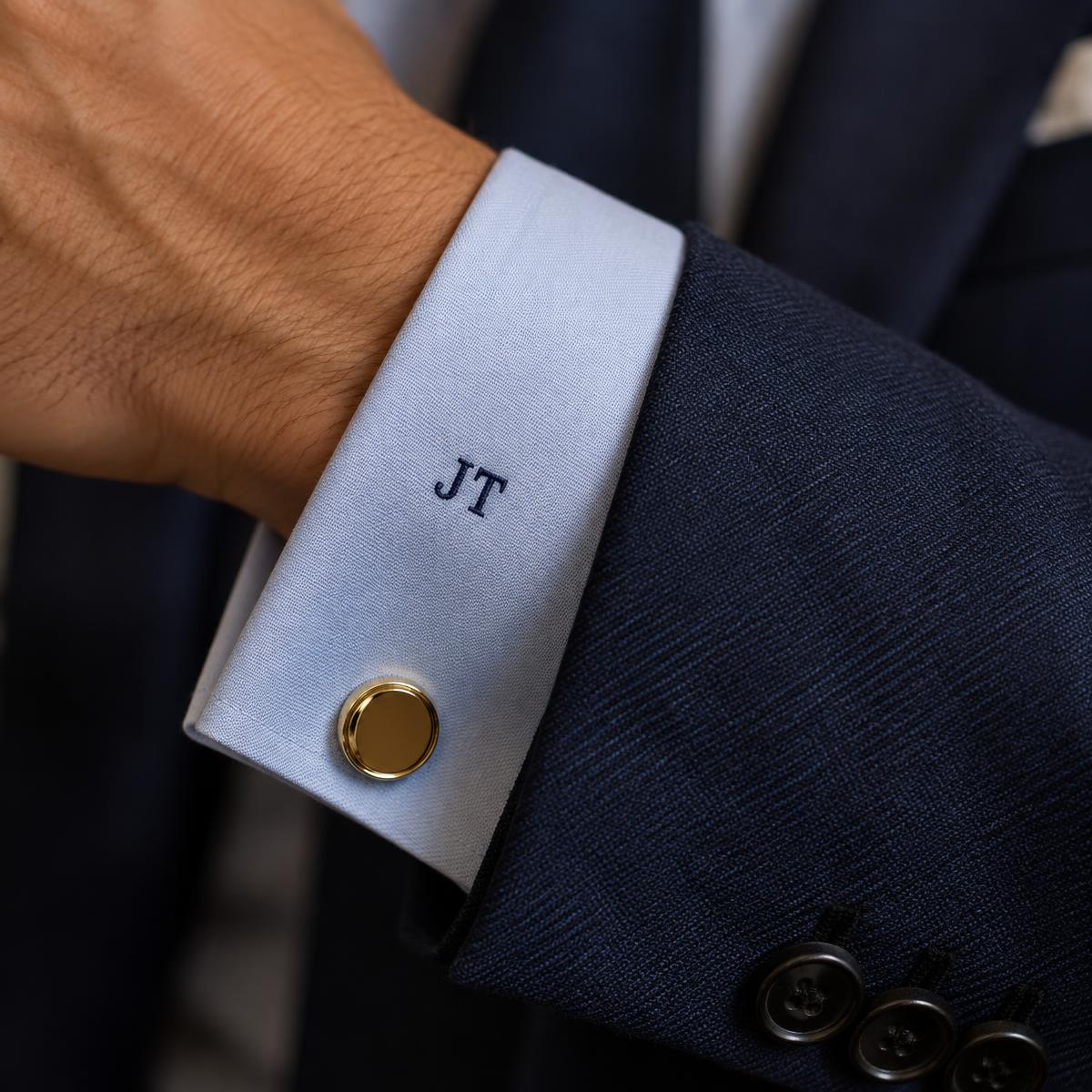 Navy embroidered JT monogram on a pale blue shirt cuff with a gold cufflink, under a navy jacket sleeve